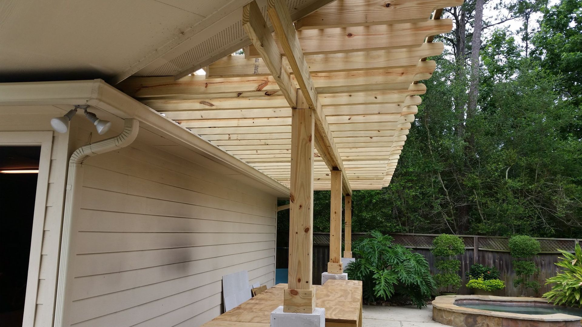 Pergola attached to a house with wooden beams and slats, supported by posts on concrete blocks.