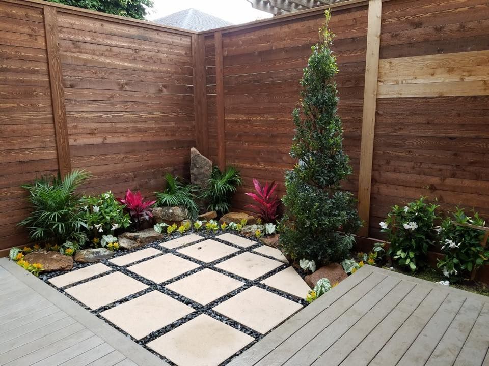 Enclosed backyard with wood fence, stone pavers, decorative plants, and a small wooden deck.