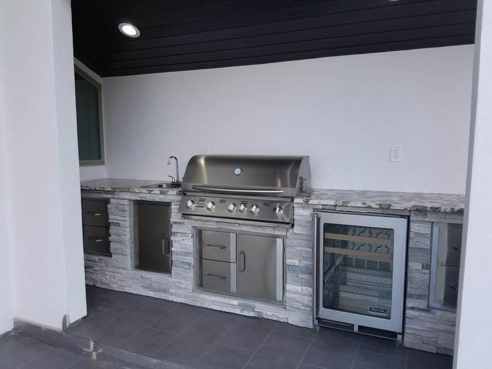 Outdoor kitchen with stainless steel grill, sink, cabinets, and wine fridge, set against a white wall with dark ceiling.