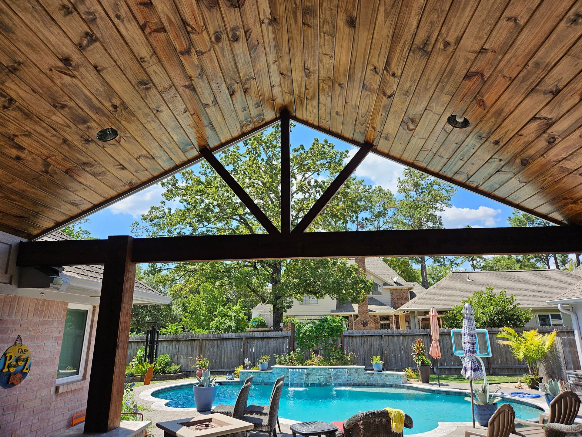 Patio with a wooden ceiling overlooking a pool, trees, and houses.