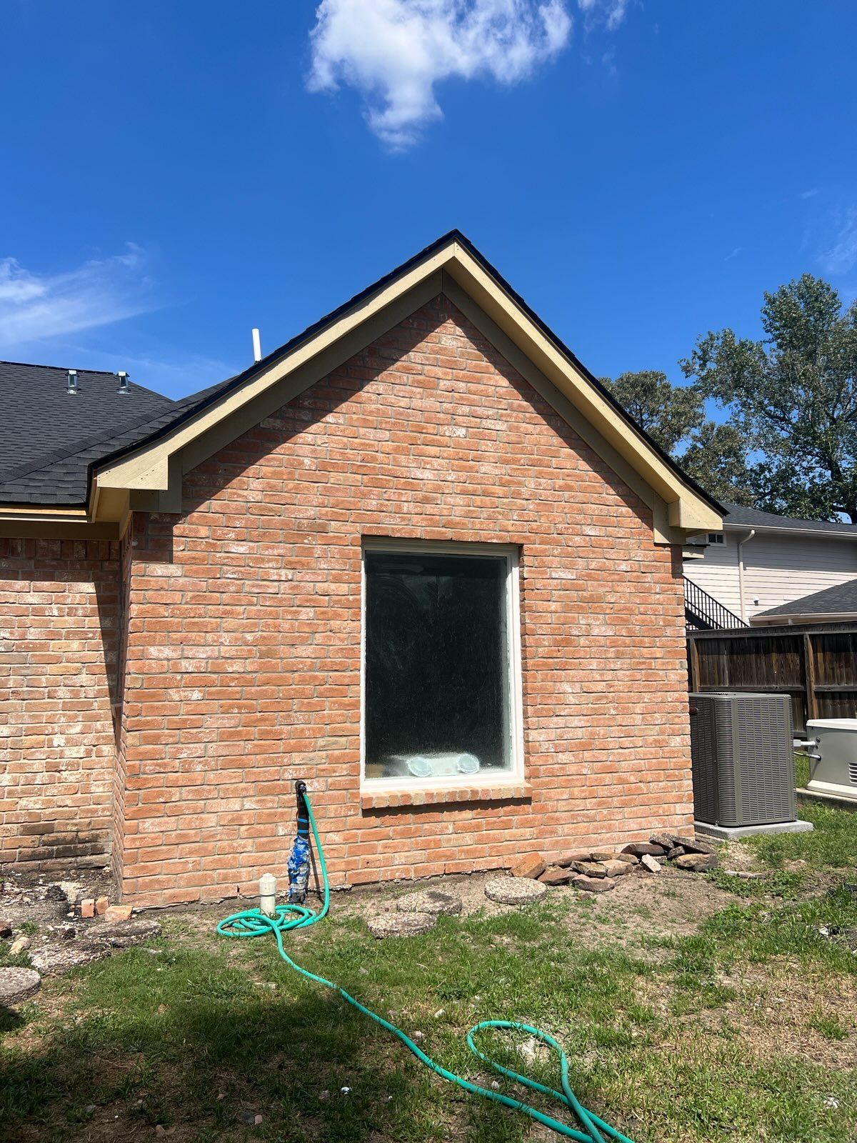 Brick wall with a window and a black roof. Green hose in the grass. Sunny day.