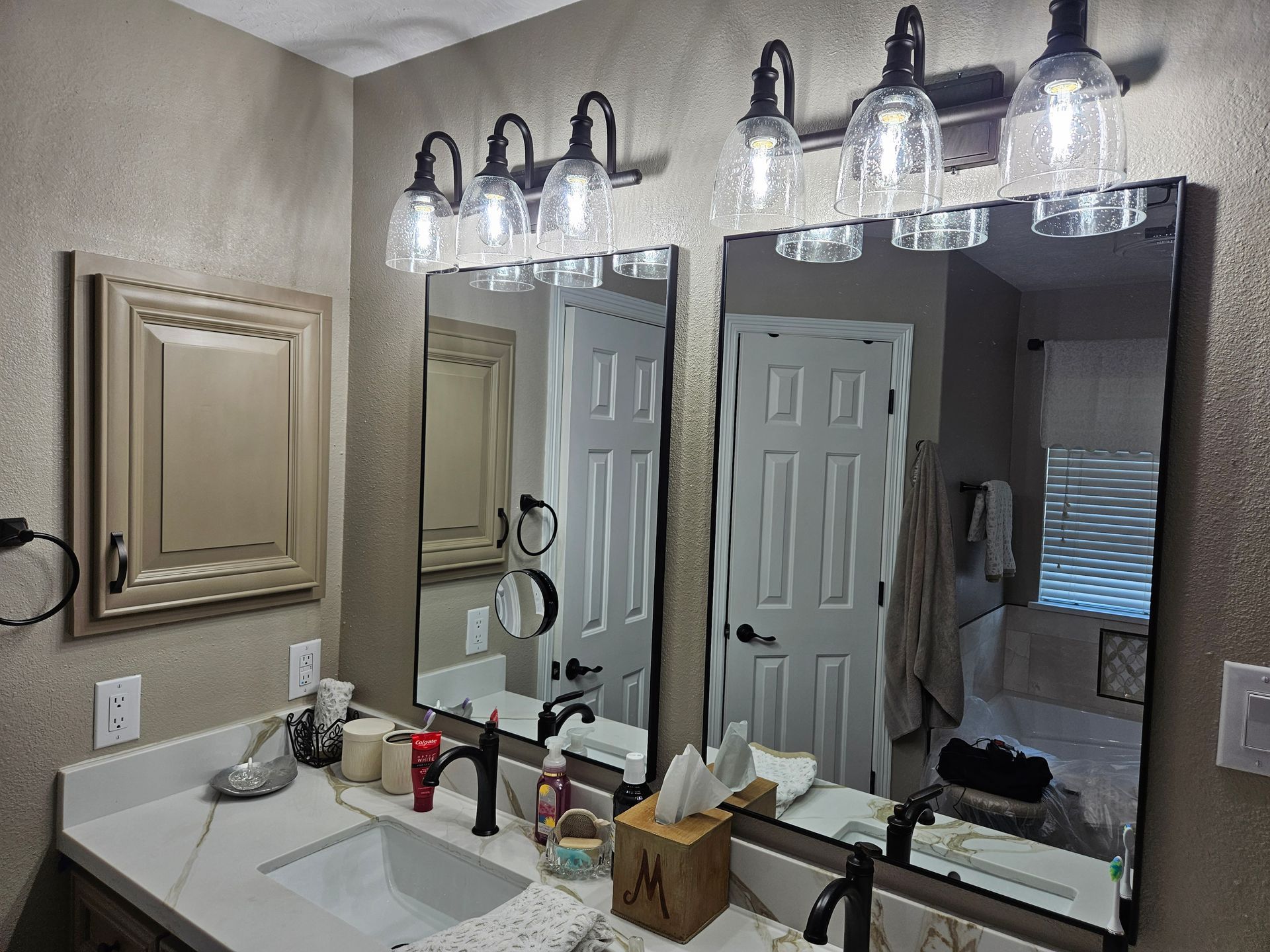 Bathroom with two mirrors, sinks, and lights. Tan walls, dark fixtures, and light-colored countertops.