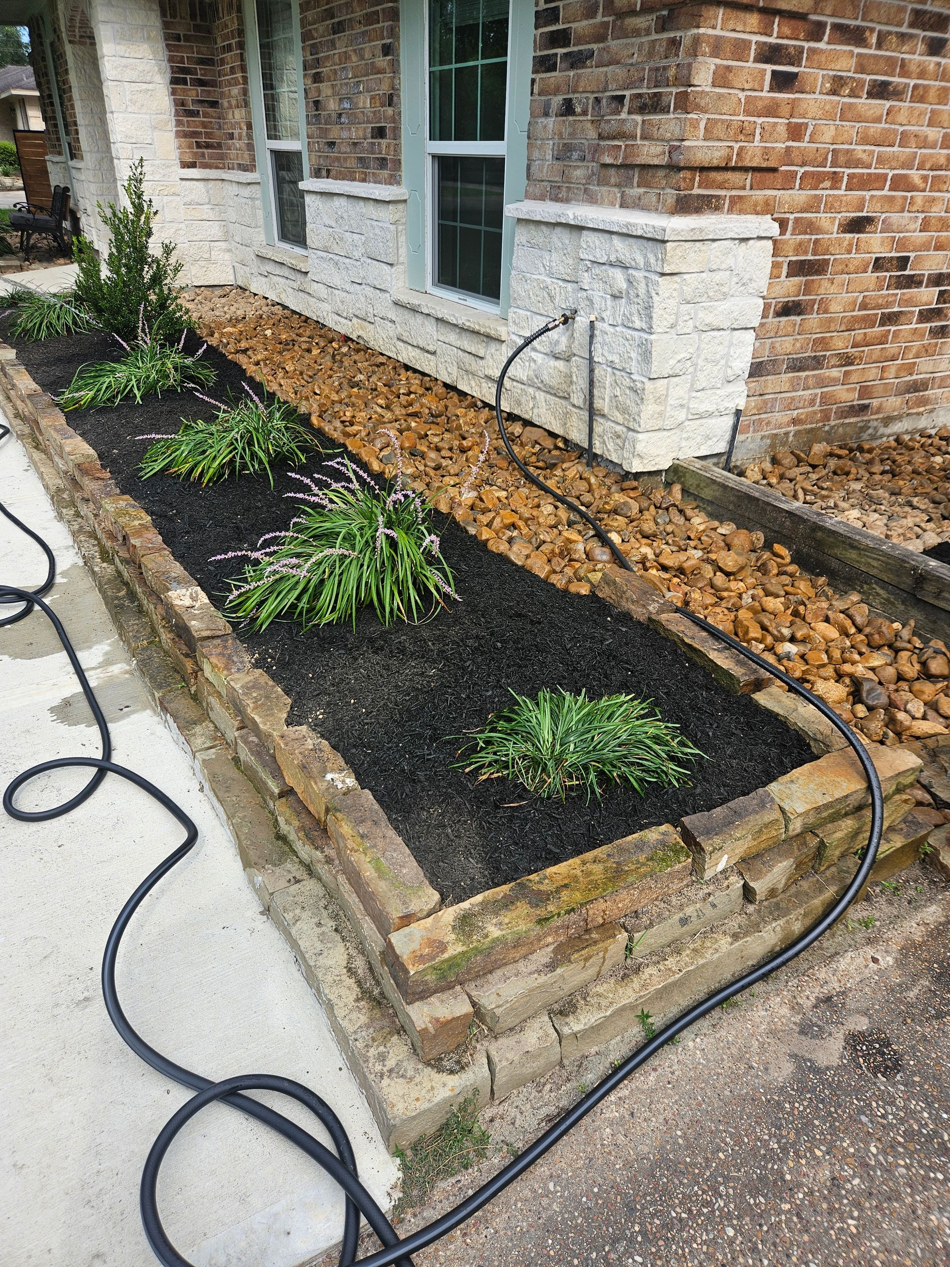Raised garden bed with plants and mulch next to a brick house with a rock border.