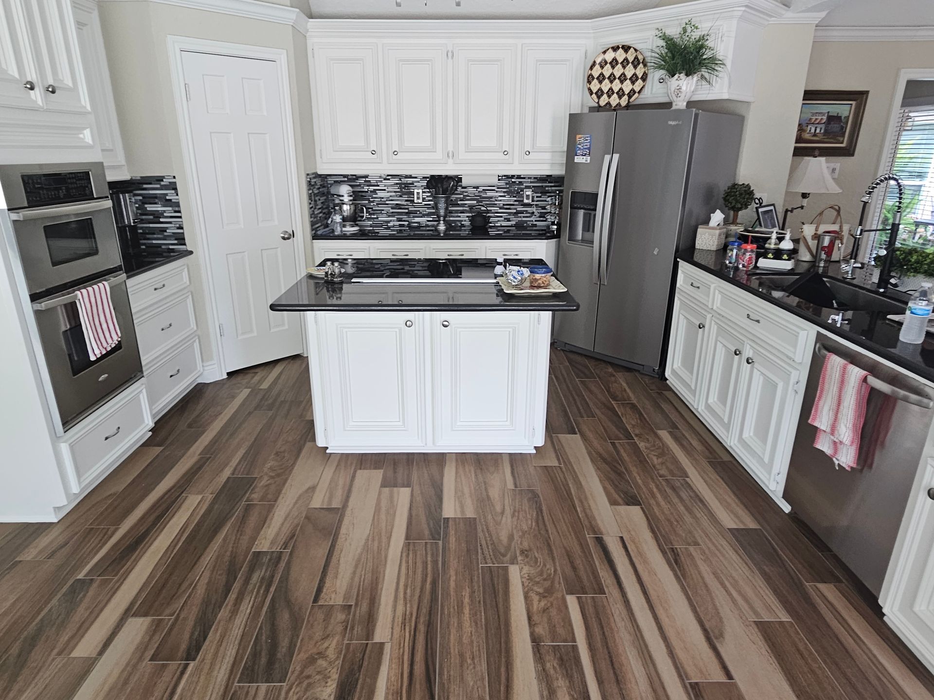 Kitchen with white cabinets, black countertops, and wood-look flooring. Island with cooktop, stainless appliances.