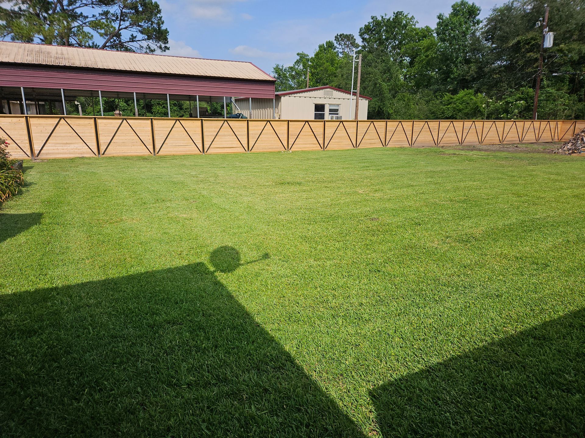Lush green lawn with a light brown wooden fence, a building, and trees in the background under a sunny sky.
