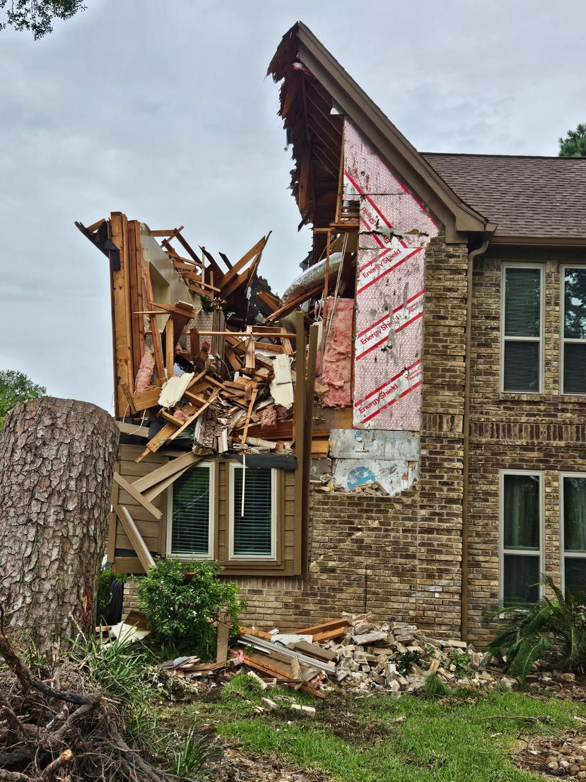House damaged by a fallen tree, exposing structure. Brown brick exterior, debris, cloudy sky.