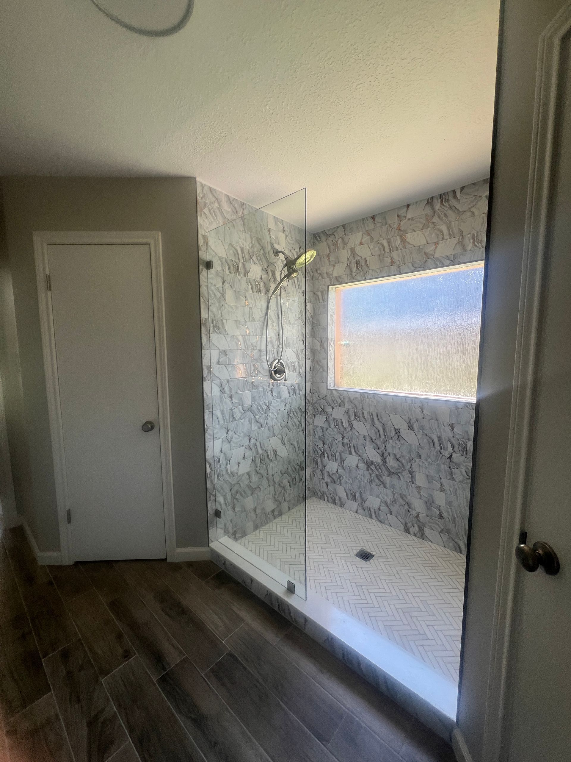 Bathroom with marble-tiled shower, glass door, and wood-look floor. A white door is on the left.