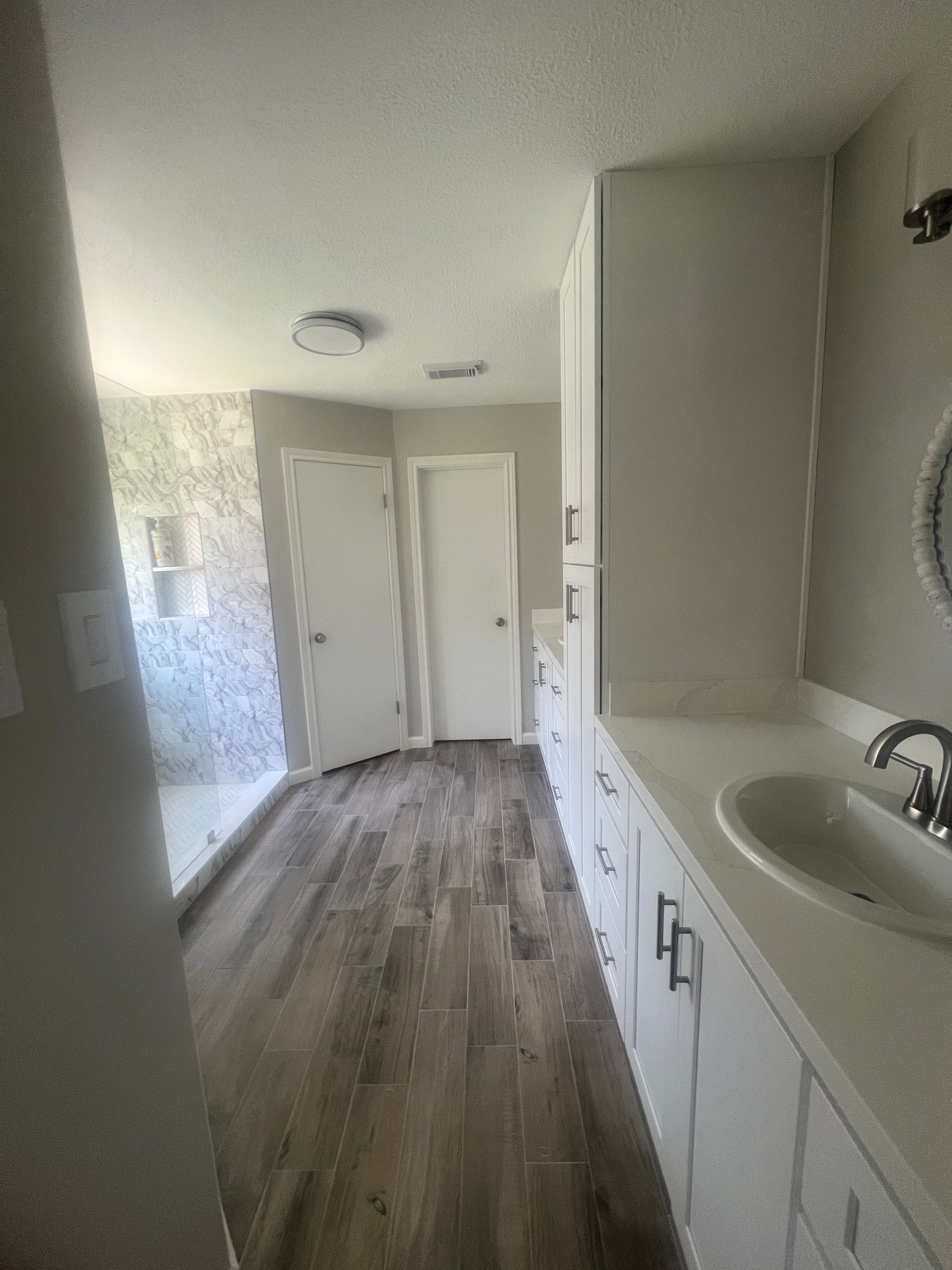 Bathroom with white cabinets, sink, and wood-look tile flooring; shower and doors in the background.