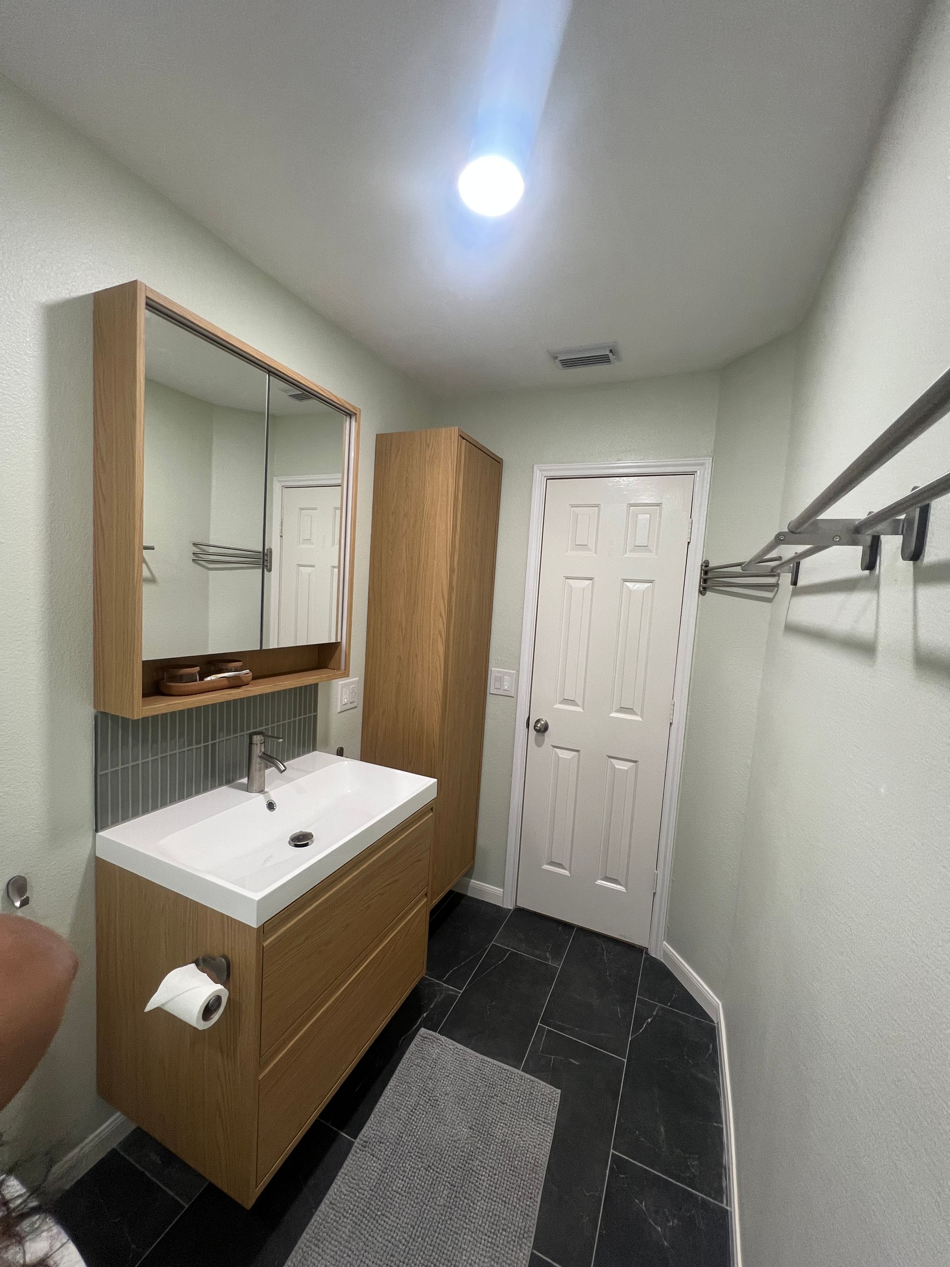 Bathroom with wooden vanity, tall cabinet, white door, black tile floor.