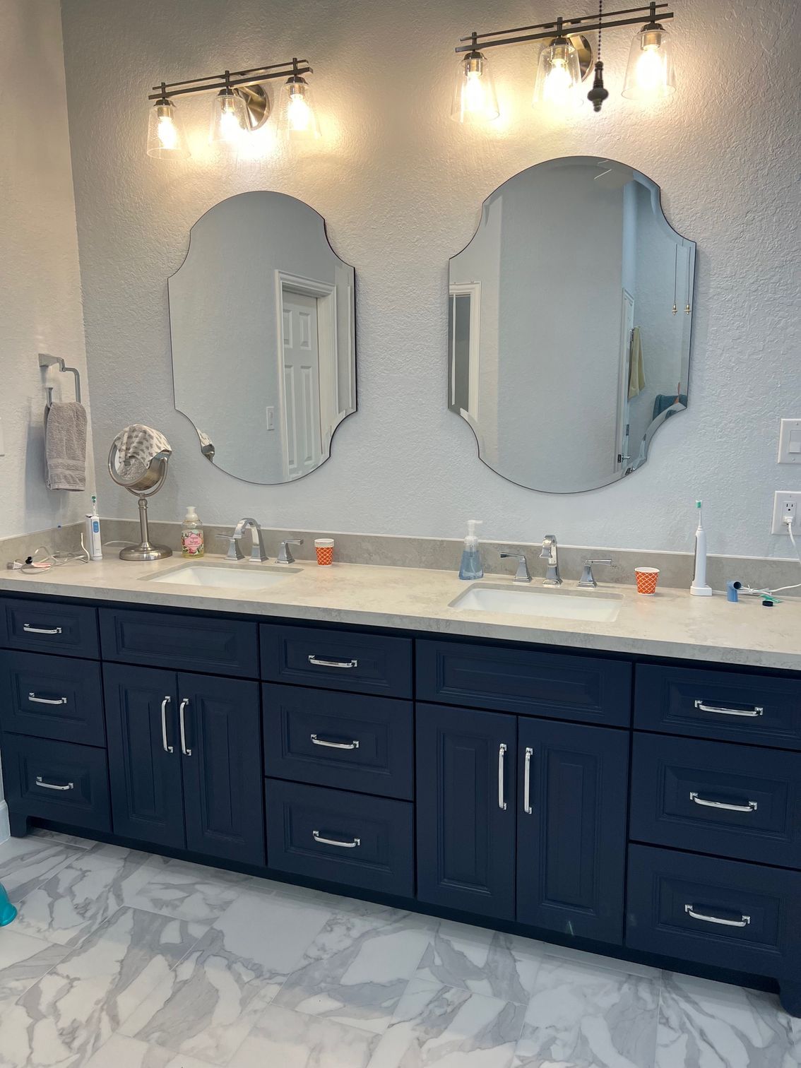 Dark blue double vanity with two ornate mirrors, two light fixtures, and marble floors in a bathroom.