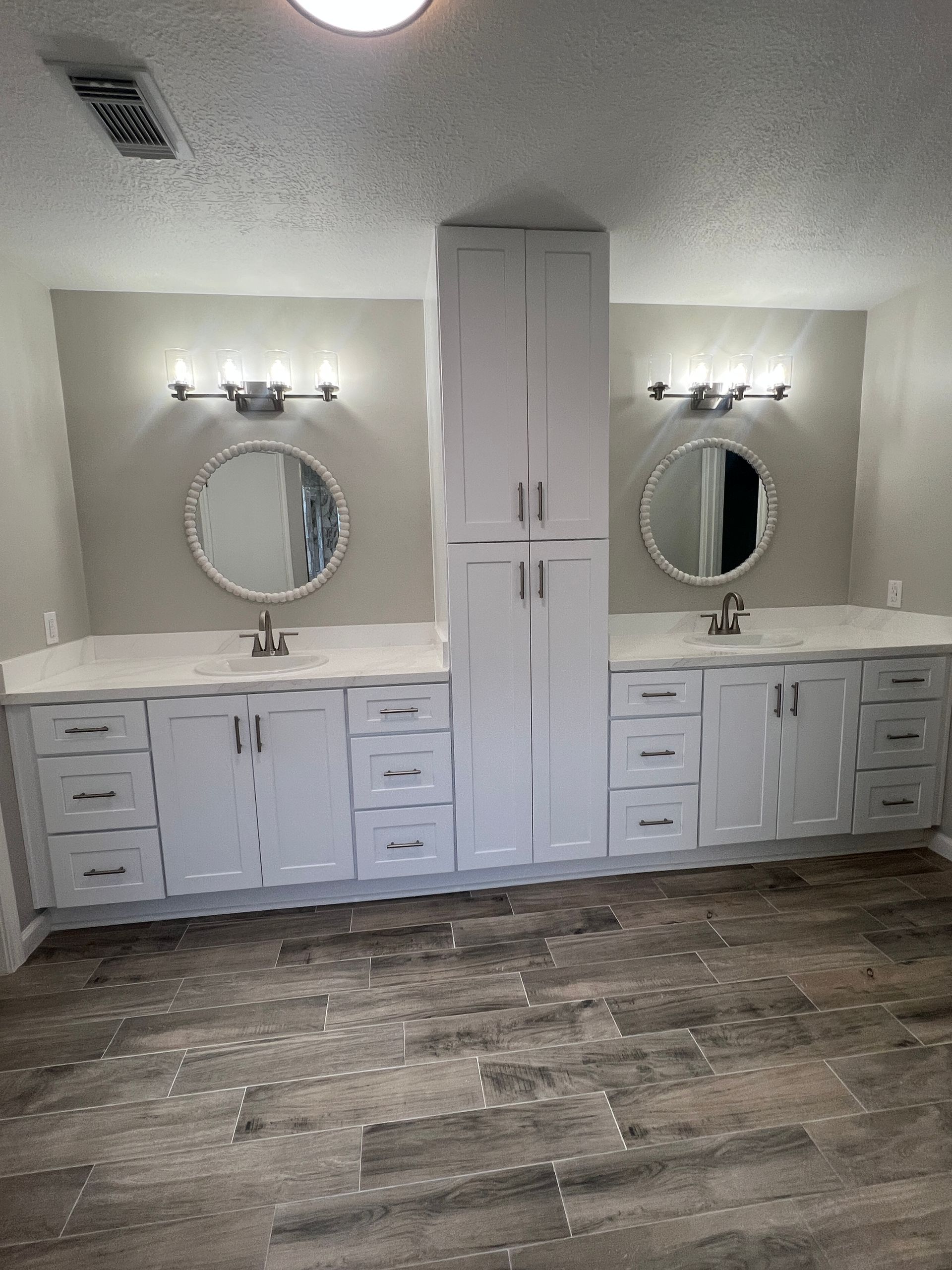 White double vanity bathroom with a tall cabinet in the middle, round mirrors, and gray flooring.