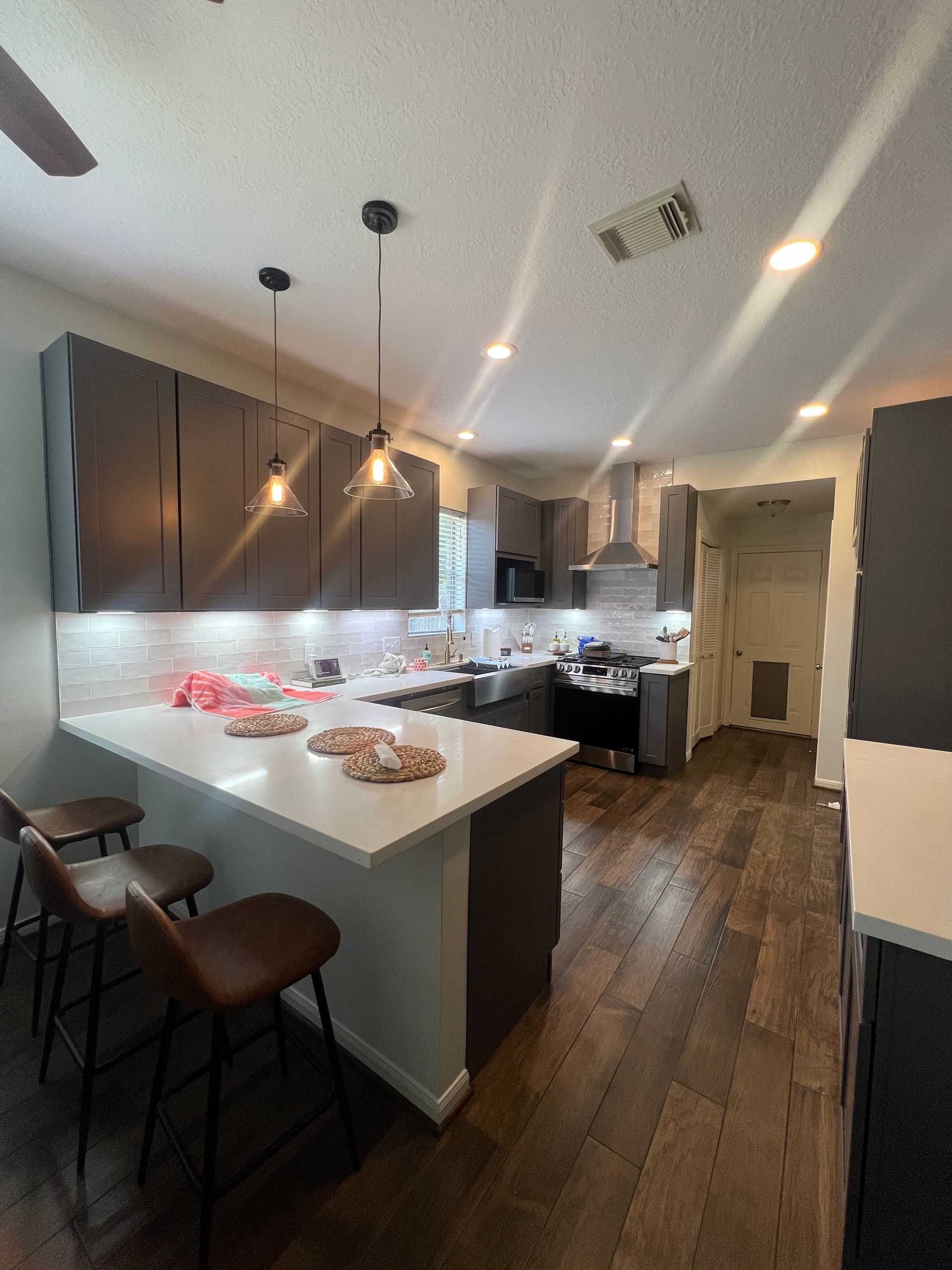 Modern kitchen with dark cabinets, white countertops, and brown wood floors. Includes a breakfast bar with stools.