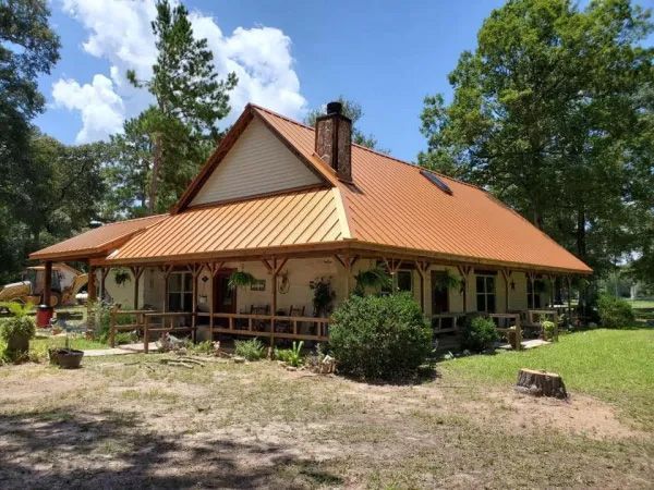 Tan house with orange metal roof, front porch, and trees under a blue sky.