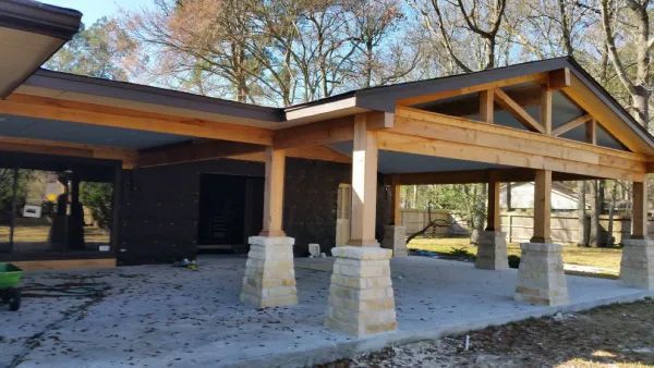 Outdoor covered patio with wooden beams and stone columns on a concrete slab, built next to a house.