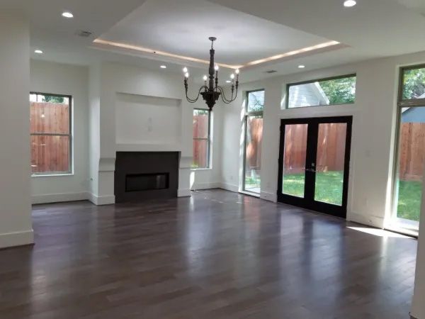 Spacious living room with dark wood floors, fireplace, chandelier, and large windows overlooking a backyard.