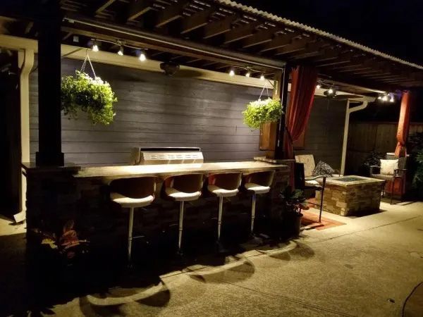 Outdoor bar at night with seating, grill, hanging plants, and string lights.