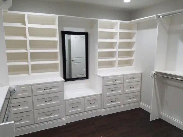 White custom closet with shelves, drawers, mirror, and bench in a room with dark wood floors.