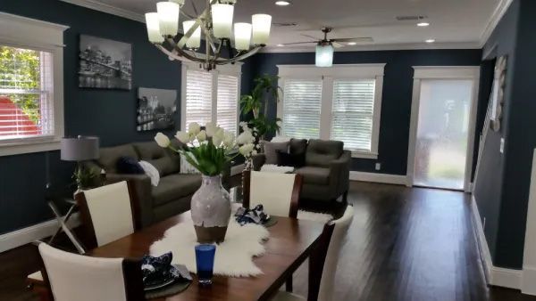 Dark-walled living room with dining table, sofas, and white trim. Dark wood floors, bright chandelier, and white flowers.
