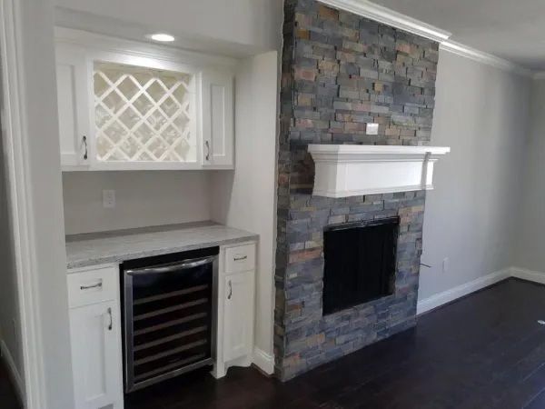 Built-in bar with wine rack and cooler, next to a stone fireplace, white cabinets, dark wood floors.