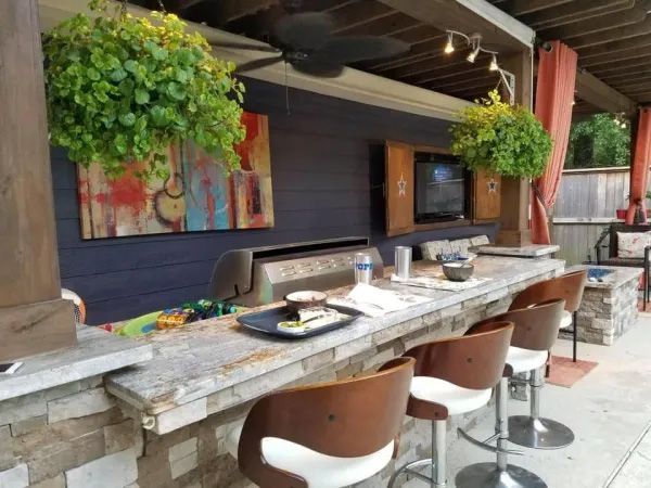 Outdoor bar with stone facade, grill, TV, and seating. Green hanging plants and colorful artwork.