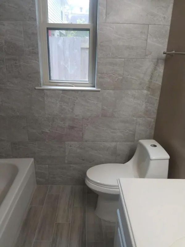 Bathroom with grey tile walls, toilet, window, and wood-look floor.