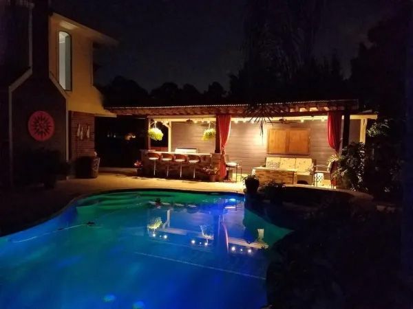 Nighttime backyard scene with a pool, bar, seating area, and overhead lighting.
