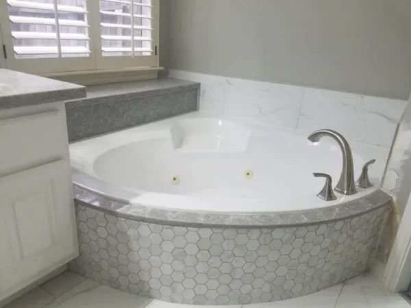 White jacuzzi tub surrounded by gray and white marble tile in a bathroom with a window.