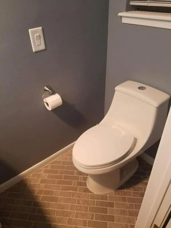 A small bathroom with a white toilet, toilet paper roll, and light switch on a gray wall.