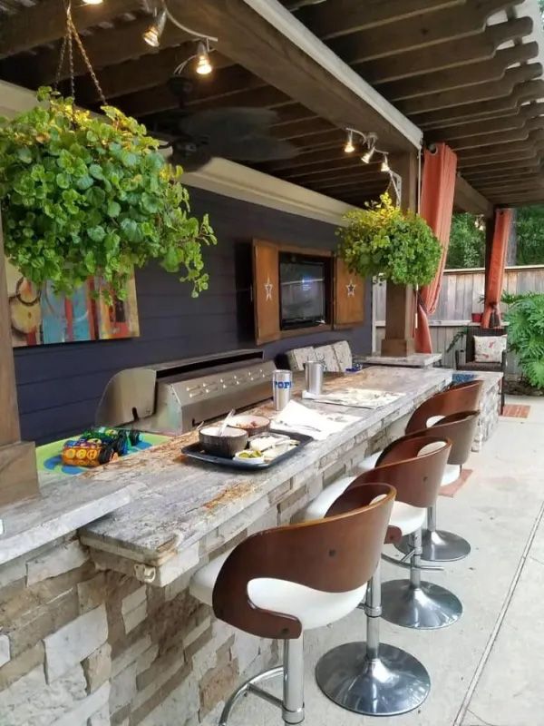 Outdoor bar with stone facade, granite countertop, and wood-backed bar stools. Hanging plants and a grill.
