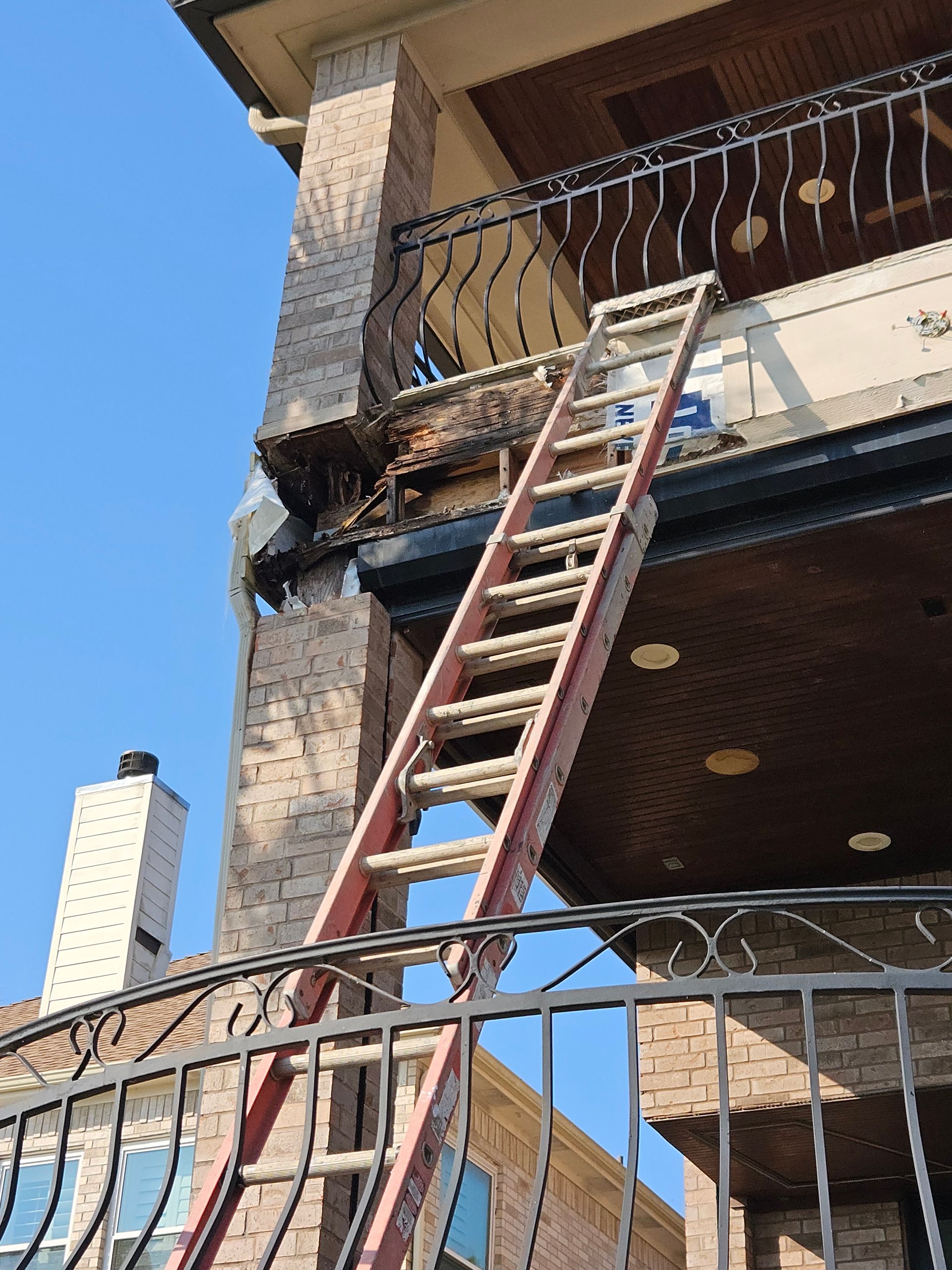 Ladder leaning against a building's damaged facade, with a wrought-iron balcony and a blue sky backdrop.