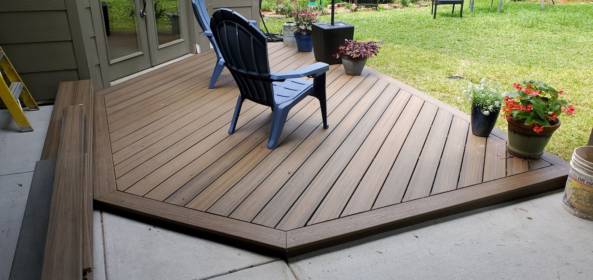 A brown composite deck with two blue chairs and potted plants sits outside a house.