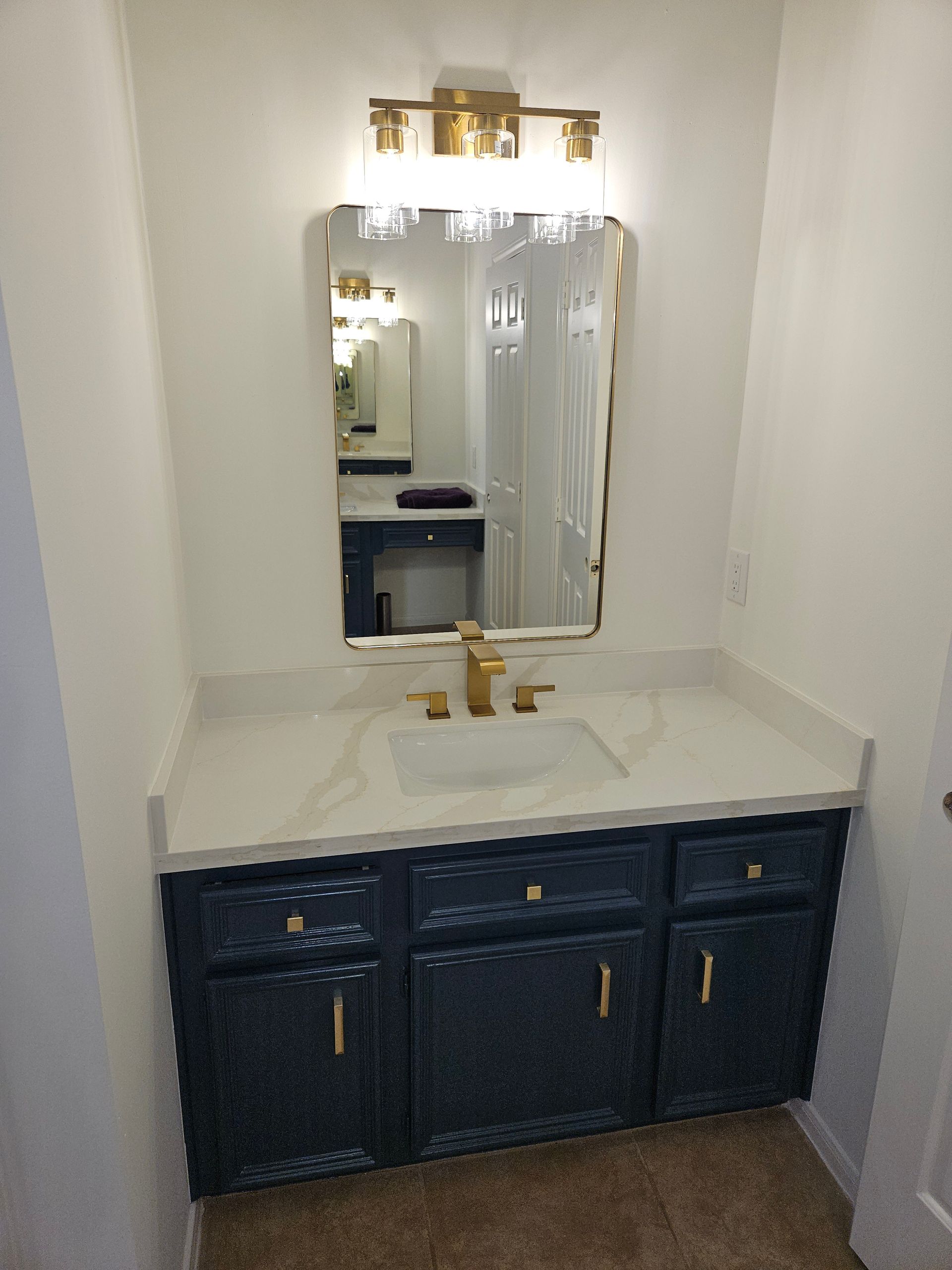 Navy blue vanity with gold fixtures and a white countertop, under a gold light fixture and mirror.