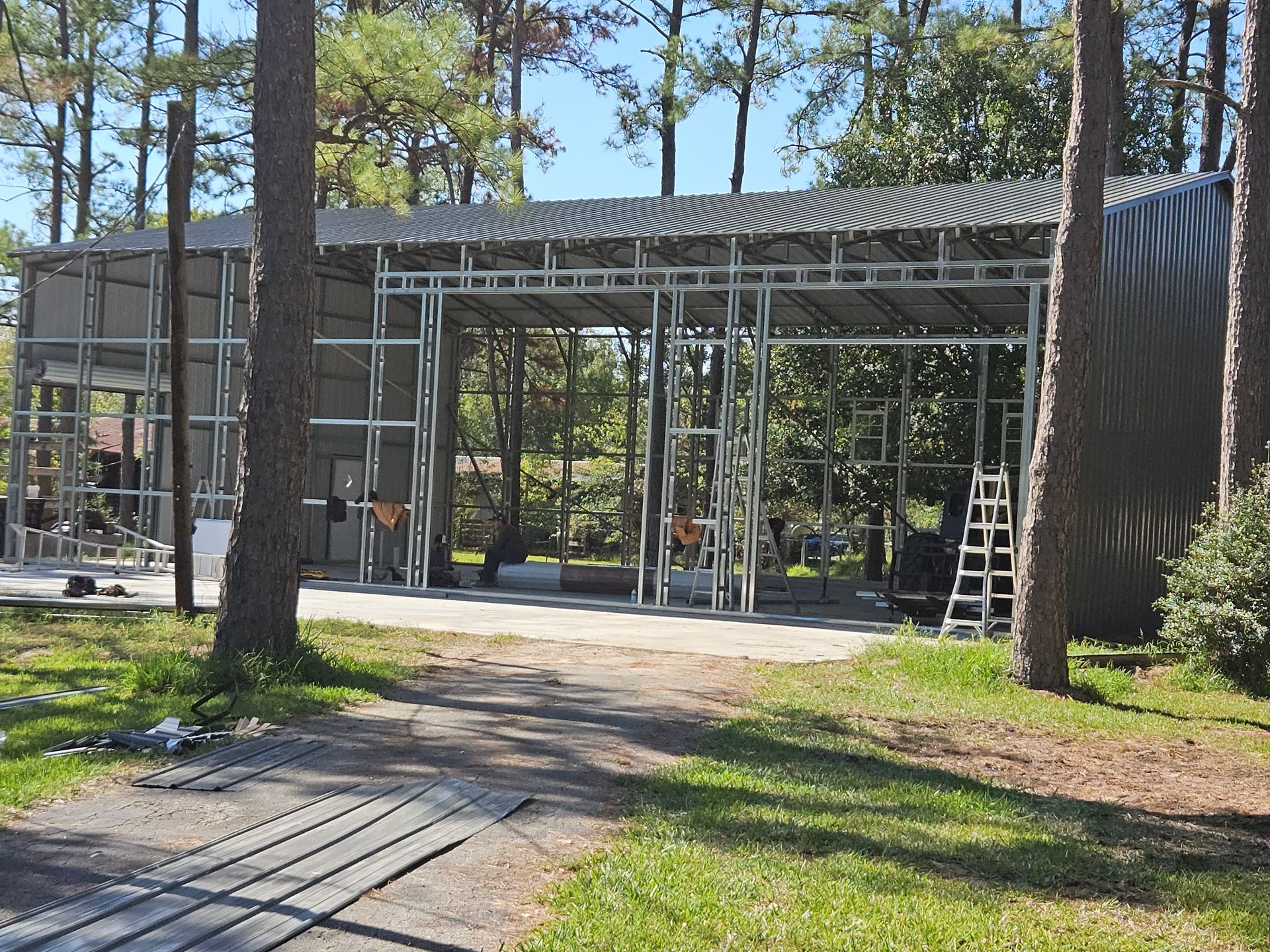 Metal frame building under construction in a wooded area; gray corrugated roof, blue sky.