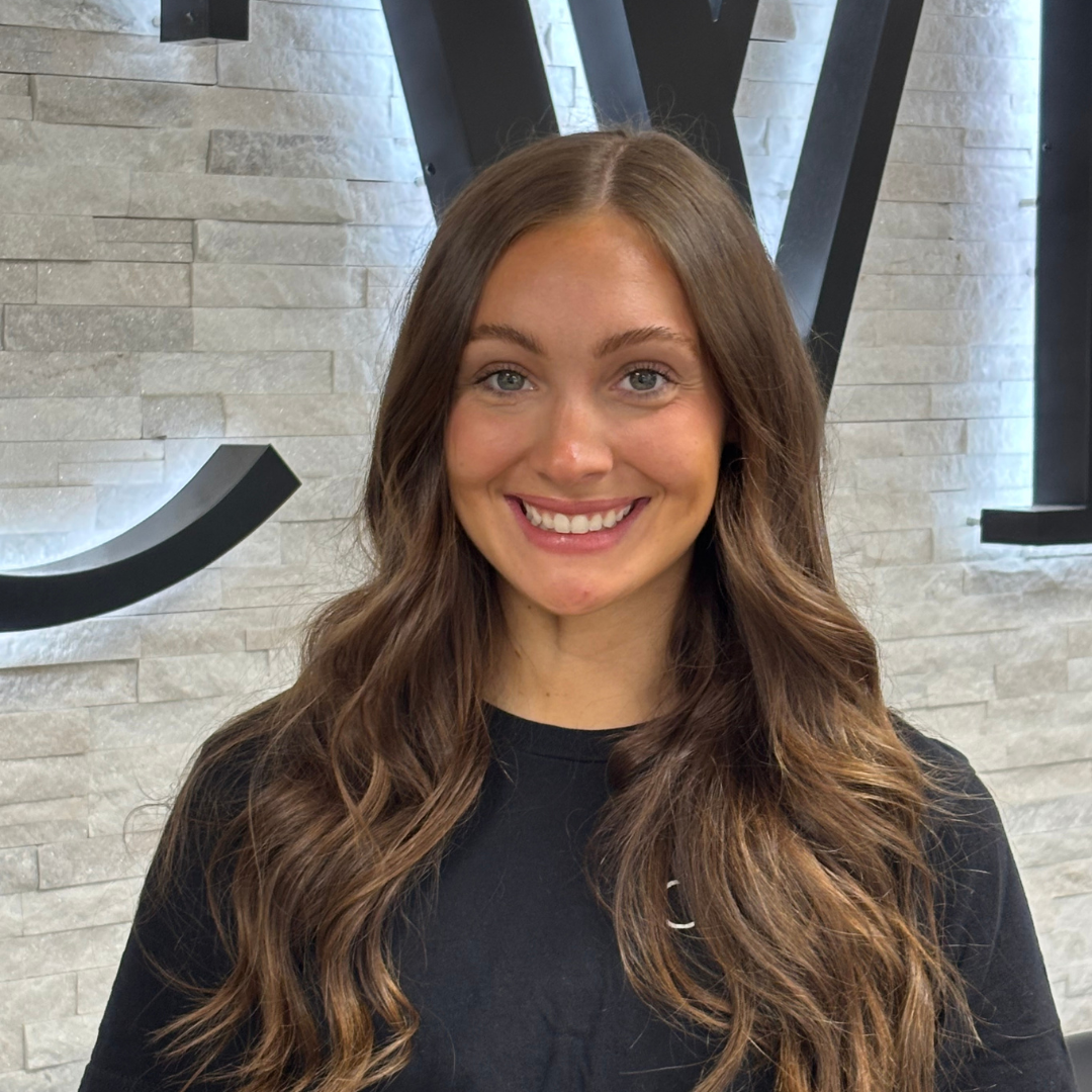 Smiling woman with long brown hair in front of a gray brick wall and large black letters
