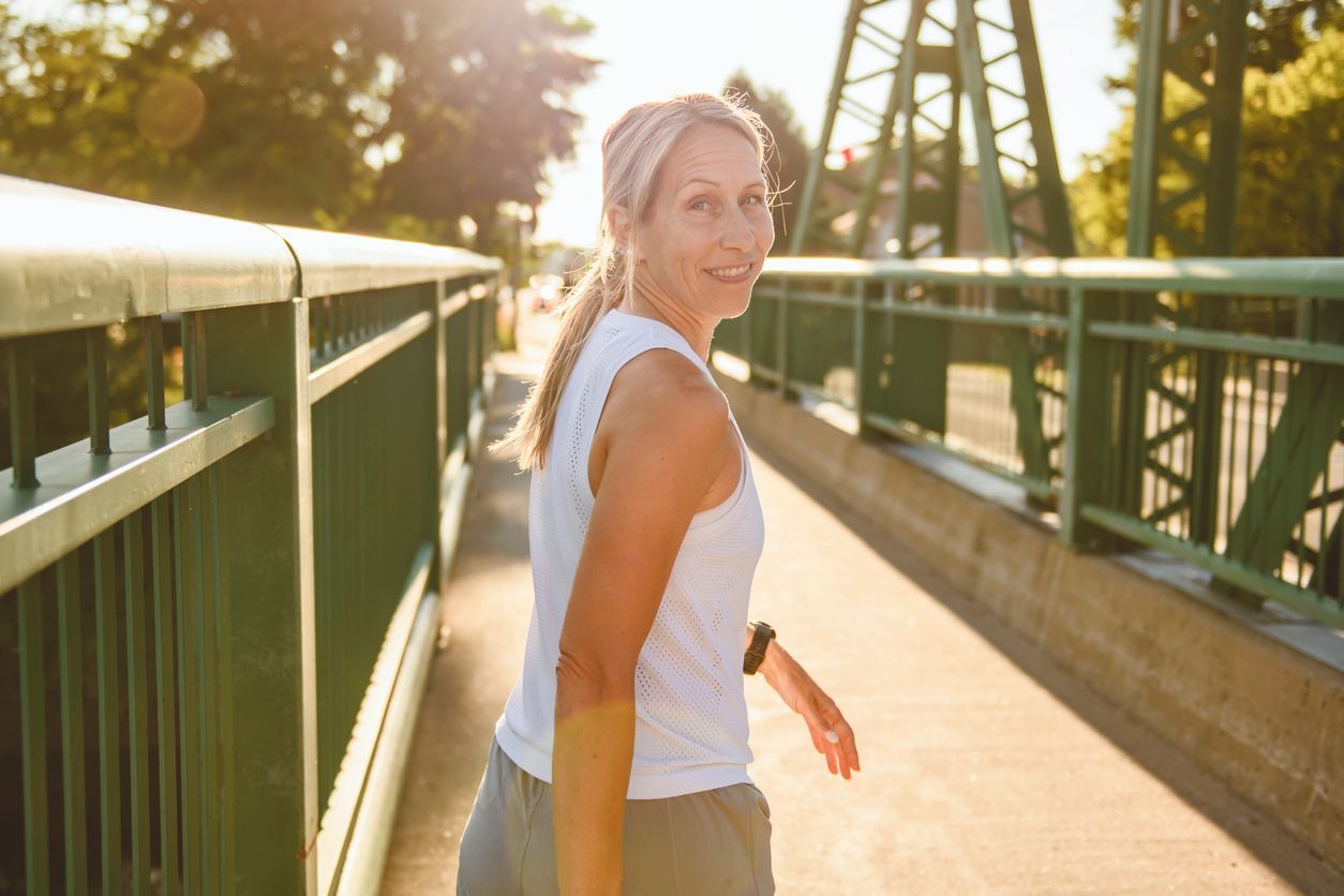 Woman walking on a sunlit bridge, turning back and smiling, with green railings and trees in the background