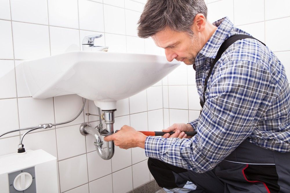 A Man Is Fixing a Sink in A Bathroom with A Wrench — Aqua Blue Plumbing Services in Bellingen, NSW
