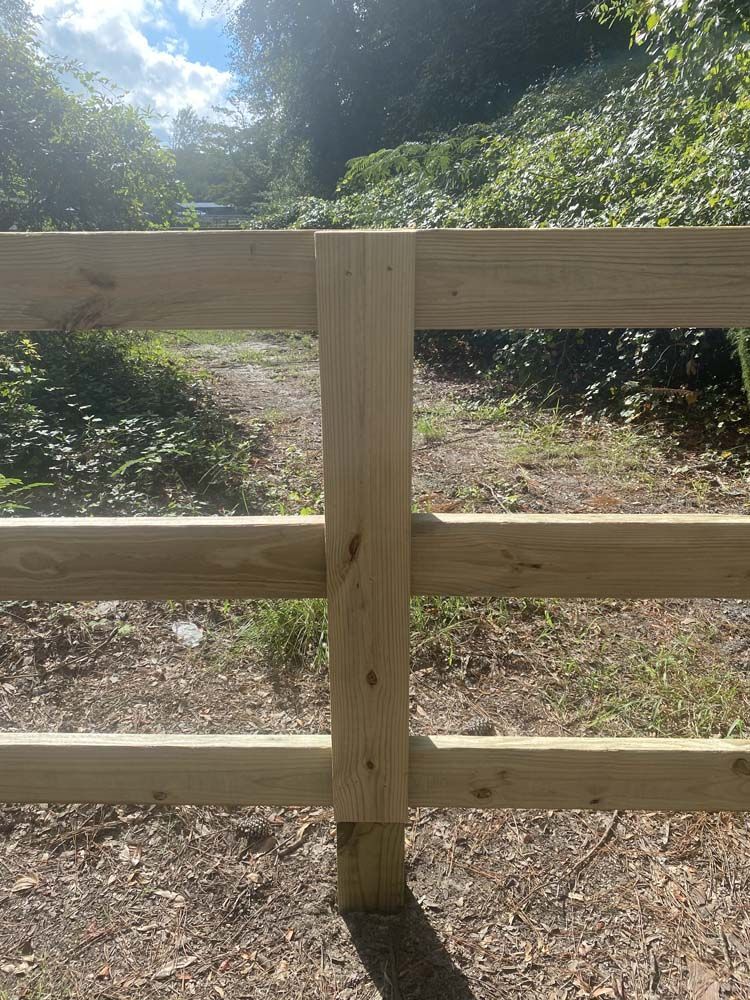 Wooden fence with three horizontal rails, a vertical post, and a path in the background.