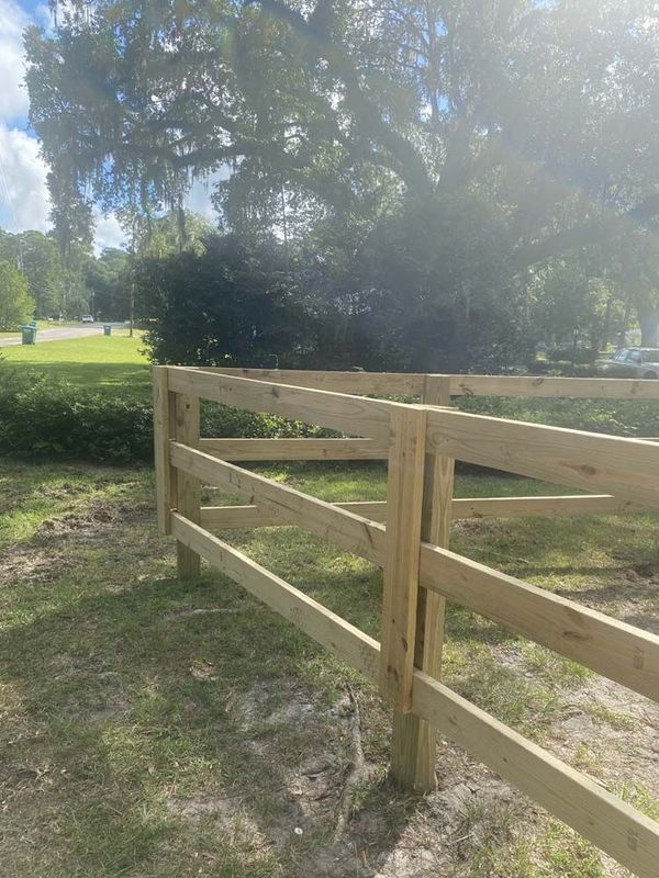 Wooden two-rail fence in a grassy yard, with a tree and road in the background.