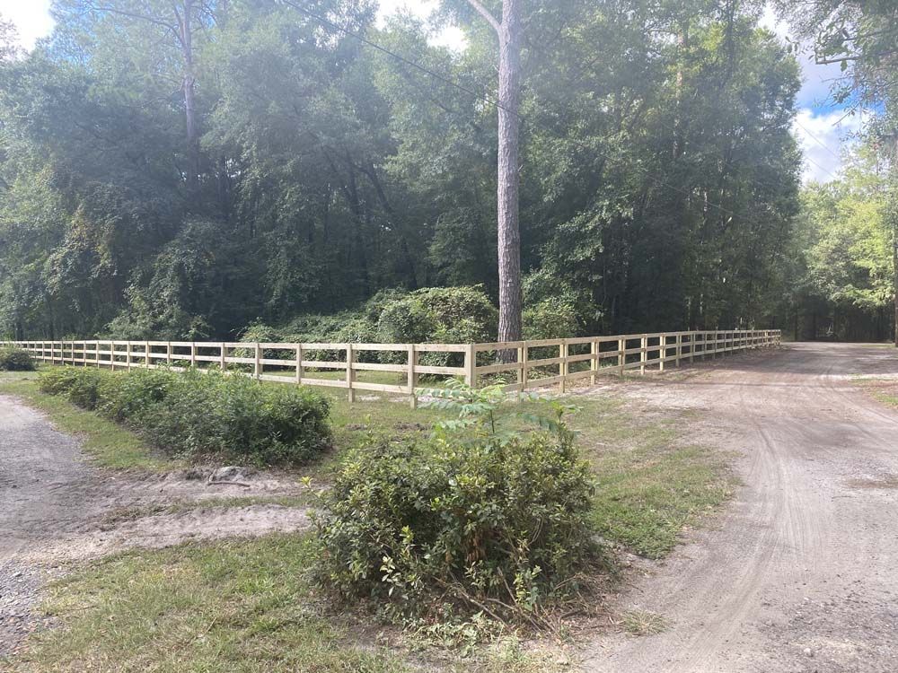 Wooden fence bordering a dirt road, next to greenery and trees under a sunny sky.