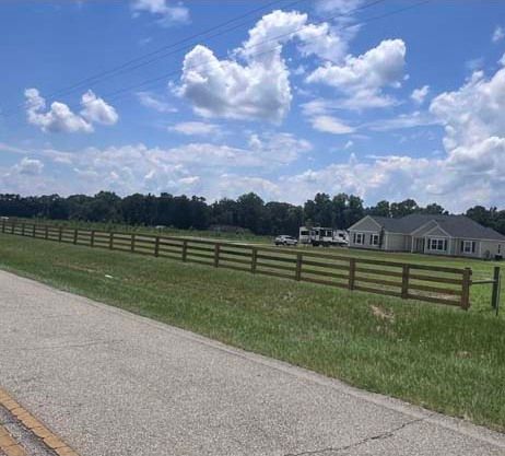 A wooden fence lines a grassy field next to a road, with homes and trees in the distance under a cloudy blue sky.