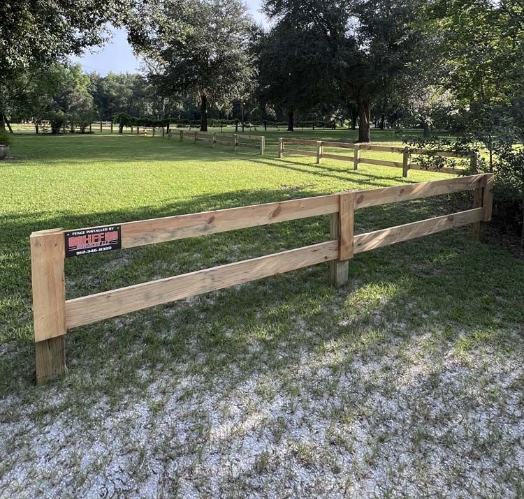 Wooden fence in a grassy field, with trees in the background under a blue sky.