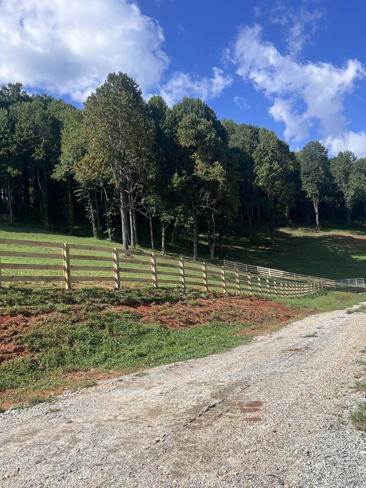 Gravel road leads to wooden fence bordering a green field and a line of trees against a blue sky with clouds.
