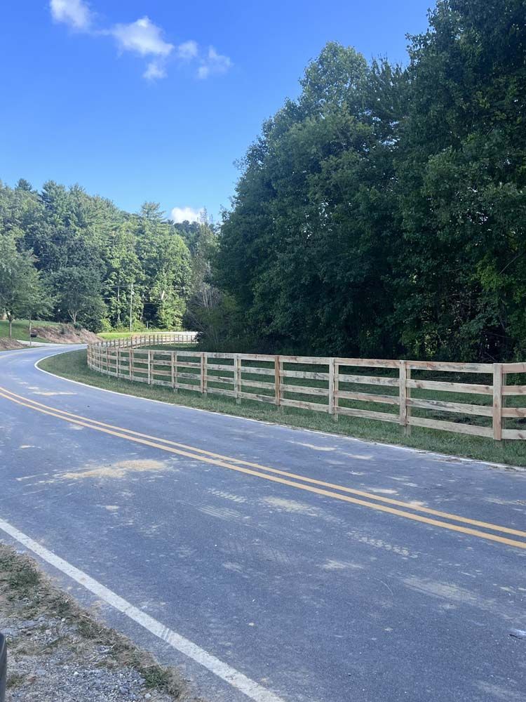 Wooden fence curves alongside a road, beside lush green trees and a blue sky.