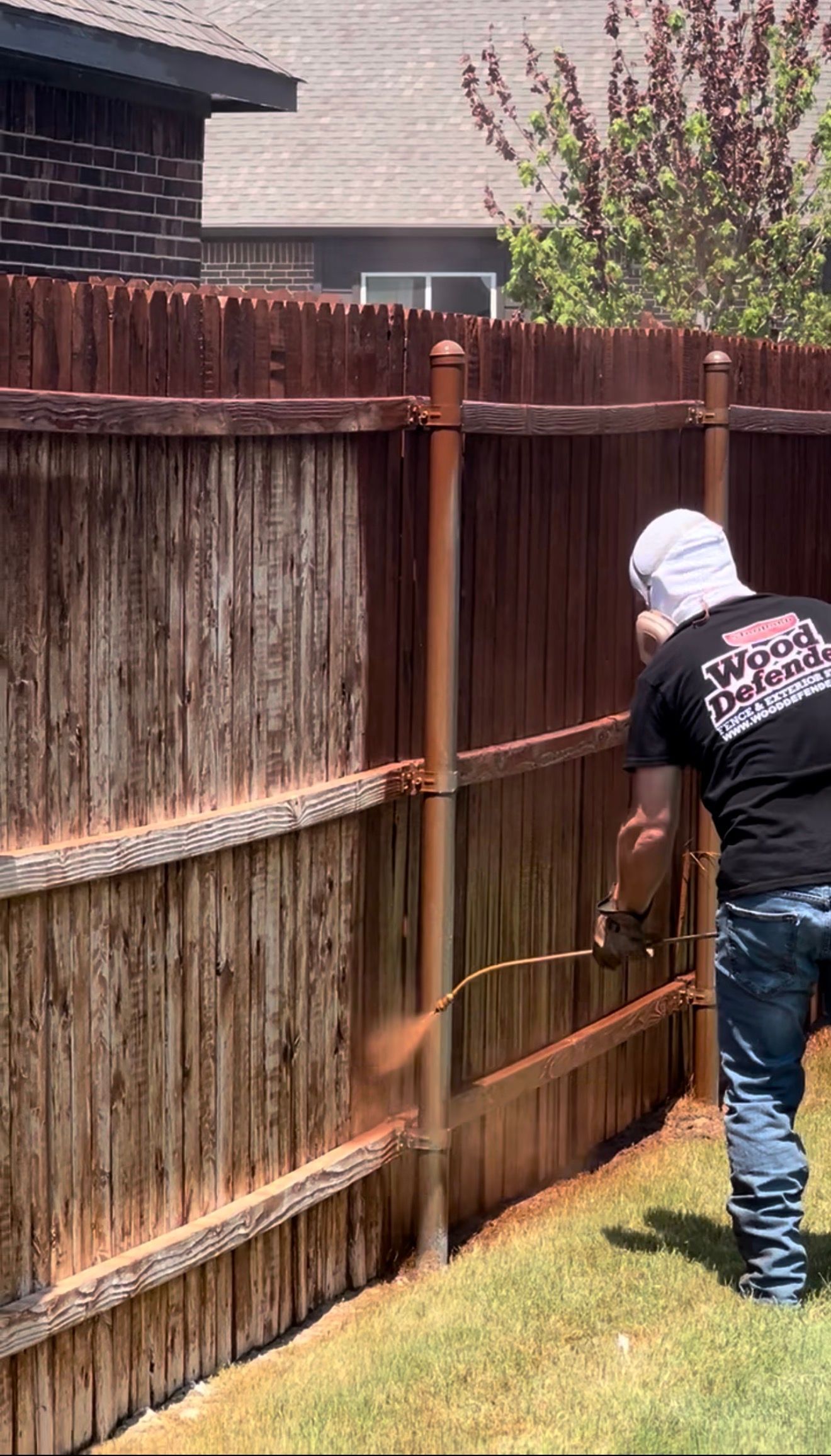 Hand painting a wooden fence with brown stain against a blue sky.