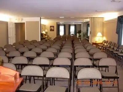 Rows of folding chairs in a beige-walled room, likely a funeral home. An empty podium is in the foreground.