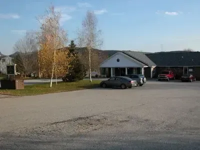 Paved parking lot in front of a low building with trees. Several cars parked on the lot.