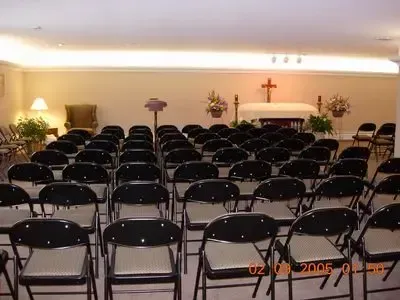 A funeral home chapel with rows of folding chairs facing a casket. A cross and flowers are present.