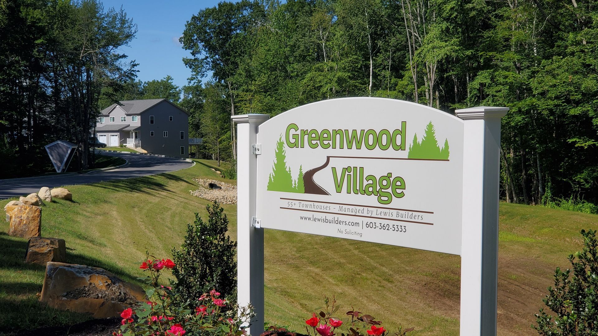 Greenwood Village sign at entrance, white and green on green lawn, with house in background.