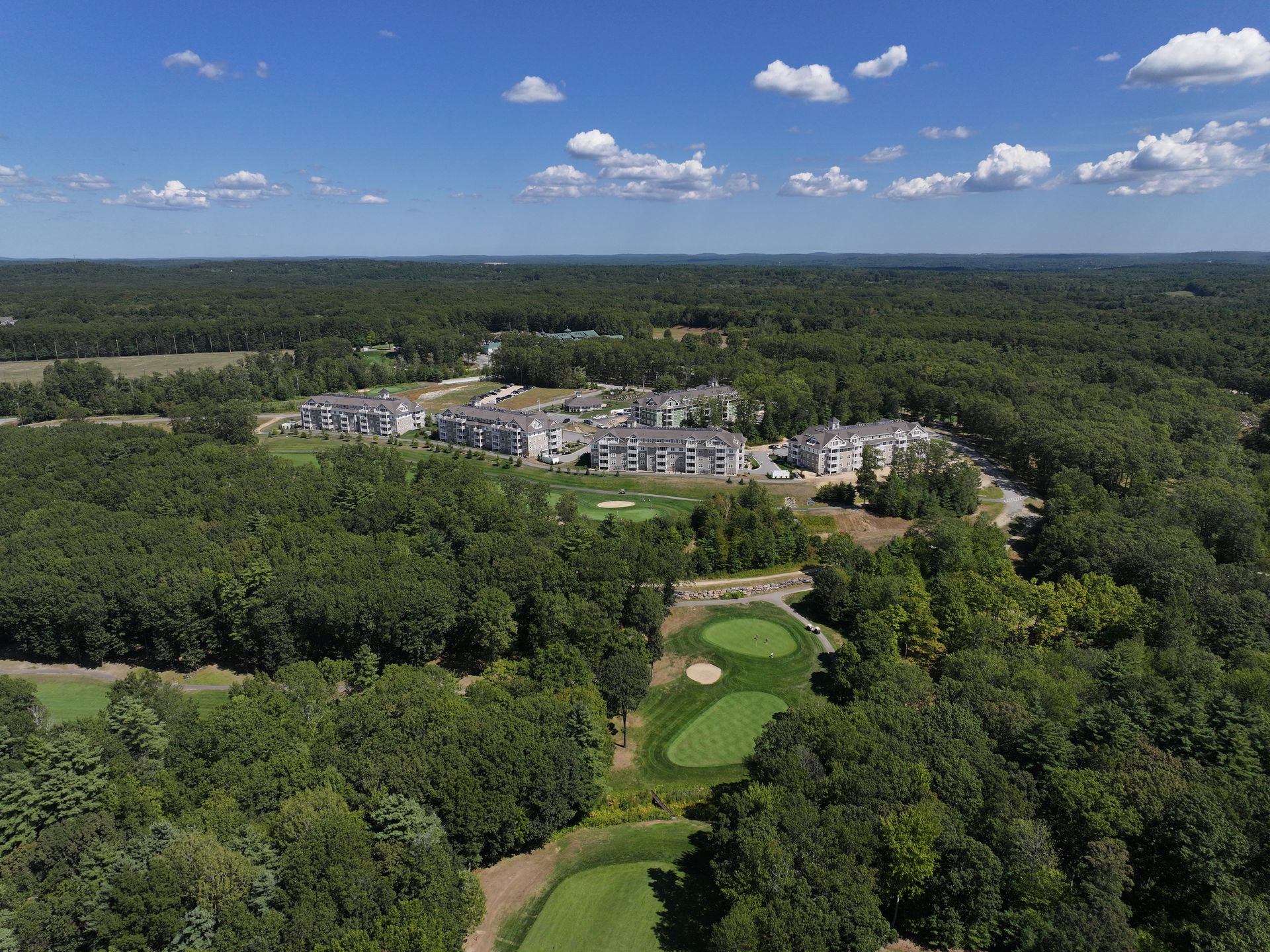 Aerial view: Apartment complex near a golf course surrounded by lush green trees under a blue sky.