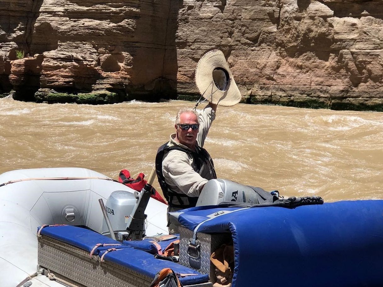 Man on a raft raises a hat in a river, canyons in the background.